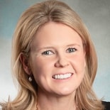 A blonde woman in a black top and pearl earrings smiles at the camera against a neutral background, reflecting medbridge professionalism.