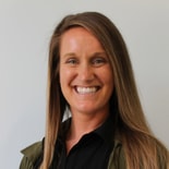 A woman with long straight brown hair and a green jacket smiles in front of a plain background, showing medbridge professionalism.