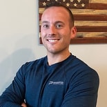 A smiling man in a blue shirt stands with arms crossed in front of American flag wall art.