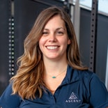 Smiling woman with long brown hair in a navy Ascent polo stands indoors by gym equipment, representing medbridge healthcare education.