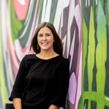 Woman with long brown hair in a black top smiles in front of a colorful mural.