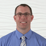 A man with short hair and glasses, wearing a blue shirt and striped tie, stands by a textured white wall contemplating medbridge's patient care software advancements.