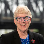 A smiling person with short light hair and blue glasses wears a black blazer with a heart-shaped pin and stands in front of a blurred bridge for medbridge.