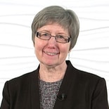 Smiling elderly woman with gray hair, glasses, and a dark blazer, set against a textured white background.