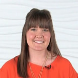 A person with long brown hair and bangs smiles, wearing an orange top with a microphone, possibly discussing medbridge innovations.