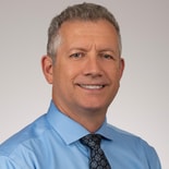 A middle-aged man in a light blue shirt and tie smiles at the camera, representing medbridge’s healthcare training.
