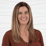 A person with long hair, smiling in a brown blouse, stands against a white textured wall, embodying Medbridge's healthcare training warmth.