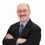 Man with glasses and a short beard in a black suit, blue shirt, and patterned tie smiling with crossed arms; medbridge style.