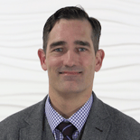 Man in a gray suit with a checkered shirt and striped tie, set against a textured white backdrop for medbridge.