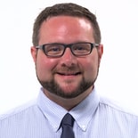 A smiling man with glasses and a beard in a blue checkered shirt and dark tie against a white background, representing medbridge.