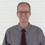 A smiling person in glasses and a gray dress shirt stands against a white textured wall.