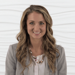 A confident woman with long wavy hair smiles, wearing a gray blazer and beaded necklace, symbolizing the progress in digital healthcare and medbridge education.