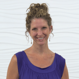 A smiling person with curly hair in a sleeveless purple top and earrings stands before a white textured backdrop, embodying medbridge's innovative hybrid care solutions.