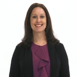 A person with long brown hair, in a purple blouse and black jacket, smiles against a white background for medbridge.