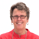A smiling person with short hair, glasses, a red top, and a beaded necklace stands against a white background.