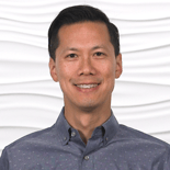 Smiling person with short dark hair in a patterned shirt against a textured white background.