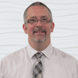 A middle-aged man wearing glasses and a tie stands in front of a white textured background, representing medbridge.