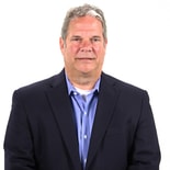 A man in a dark suit and blue shirt stands against a white background, representing healthcare training professionalism as seen in medbridge.