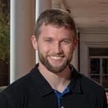 A bearded man in a black shirt smiles confidently in a covered walkway, contemplating hybrid care's impact on healthcare.