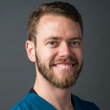 A bearded man in a teal shirt smiles confidently, suggesting expertise in hybrid care solutions, on a gray background.