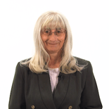 A person with long gray hair and glasses, wearing a dark blazer, stands confidently against a white background.