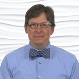 Individual with glasses, wearing a blue shirt and checkered bow tie, stands against a white textured background for medbridge.