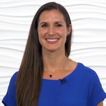 A woman with long brown hair in a blue blouse and necklace smiles against a textured white background, representing medbridge's healthcare training.