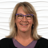 A confident, smiling woman in a purple top and black cardigan stands against a wavy white background, representing medbridge's blend of digital healthcare and traditional practices.