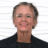Older adult with glasses and a blazer, smiling against a light background.