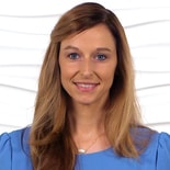A smiling woman in a light blue blouse stands against a textured white wall, representing Medbridge's digital healthcare innovation.