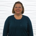 A person with shoulder-length hair in a dark teal sweater stands against a white textured wall, representing Medbridge's passion for healthcare education.
