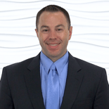 A confident man in a dark suit and blue tie poses against a white background, representing healthcare expertise for medbridge.