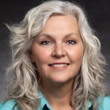 A smiling person with long, wavy gray hair in a turquoise top against a dark background represents the innovative spirit of digital healthcare by medbridge.