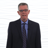 A man in a suit and glasses stands against a textured wall, representing medbridge's modern healthcare education with professionalism.