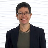 A person with short dark hair and glasses in a black blazer stands against a textured background, symbolizing healthcare education's blend into hybrid care environments for medbridge.