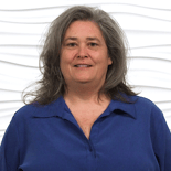 A person with shoulder-length gray hair in a blue shirt stands calmly against a white textured wall, representing medbridge expertise.