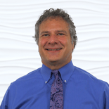 A smiling person with curly gray hair, in a blue shirt and patterned tie, represents the warmth of healthcare education at medbridge.