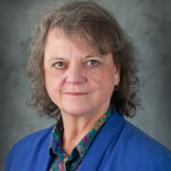 A person with shoulder-length hair in a blue blazer and paisley shirt poses professionally, embodying medbridge healthcare training.