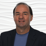 A person with short dark hair in a blue shirt and dark jacket stands against a wavy white background for medbridge.