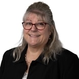 Smiling woman with long gray hair and red glasses, in business attire, stands before a white background for medbridge healthcare training.