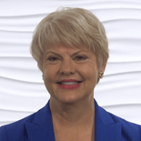 Person with short light hair in a blue blazer smiles at the camera against a white textured background.