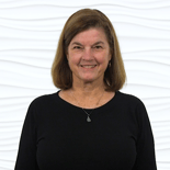 A person with shoulder-length hair, wearing a black shirt, smiles in front of a white textured wall, passionate about digital healthcare innovations by medbridge.