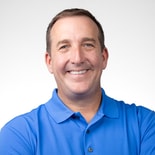 Man with short brown hair in a blue polo shirt smiles with arms crossed, standing against a white background for medbridge digital healthcare.
