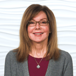 A smiling person with glasses and brown hair wears a gray sweater over a red top, representing Medbridge in digital healthcare.