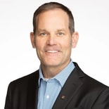 A man in a black suit jacket and blue shirt smiles at the camera, representing the future of digital healthcare at medbridge.