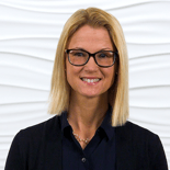 A smiling person with blonde hair and black glasses wears a dark shirt and necklace against a white textured background, promoting medbridge.