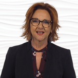 A person with wavy auburn hair and glasses speaks while wearing a dark blazer and floral blouse, set against a textured white background.