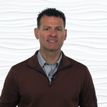 A person with short curly hair in a brown pullover stands against a textured white background, representing modern digital healthcare and medbridge flair.