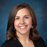 A smiling person with brown hair in a black blazer represents medbridge's hybrid care against a dark blue backdrop.