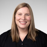 A person with straight, shoulder-length blonde hair and a black shirt smiles at the camera in front of a light gray background.